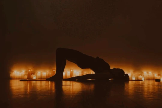 Silhouette of a person holding a bridge pose on a mat, surrounded by candles in a dimly lit room.